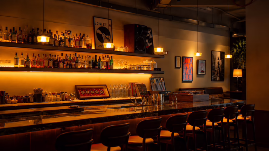 Warmly lit interior of Toma y Toma cocktail bar in Bangkok featuring marble counter seating, vinyl decor and back bar shelves.