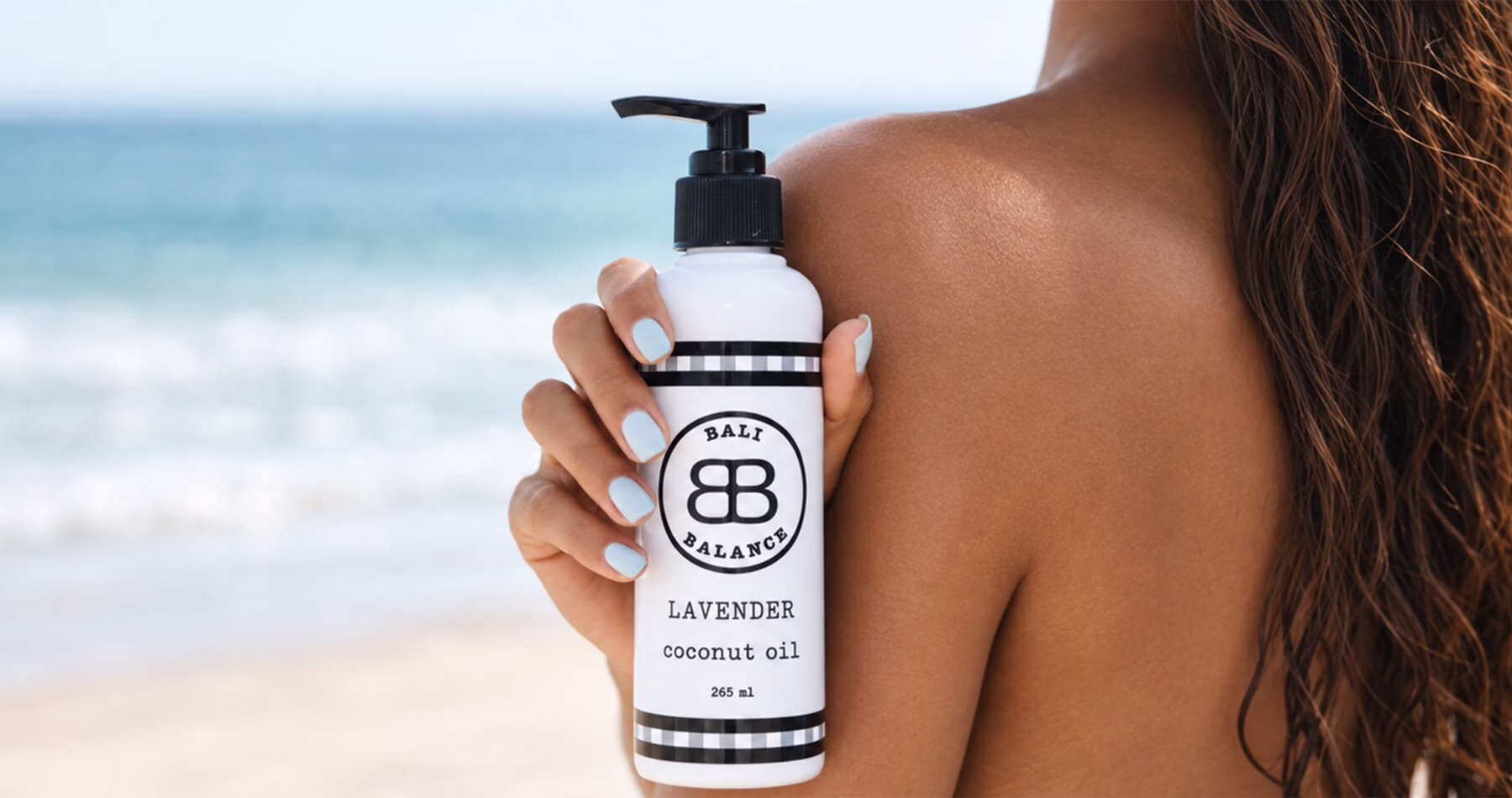A bottle of Bali Balance Lavender Coconut Oil held against a woman's shoulder, with the sea in the background.