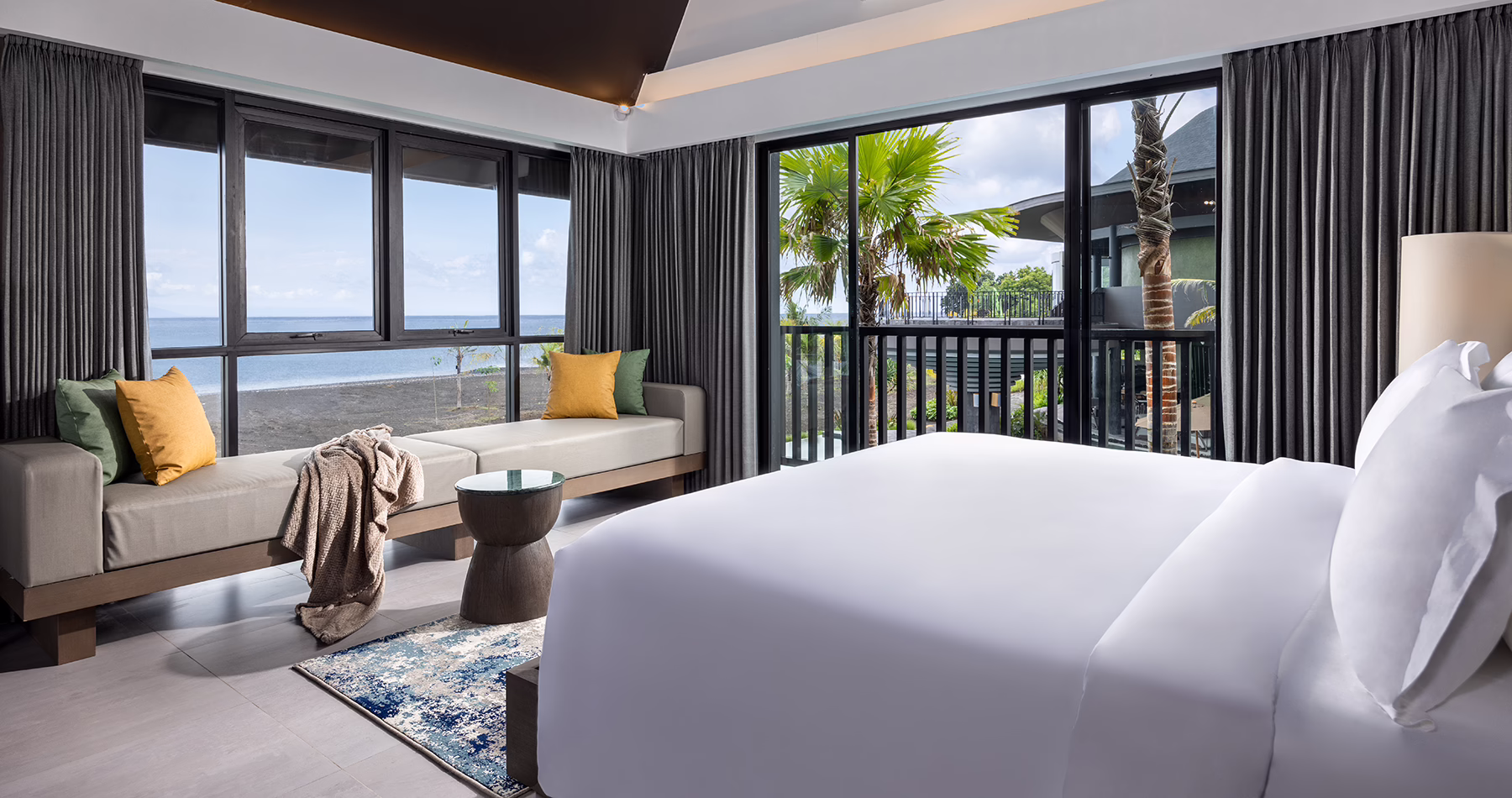 One-bedroom accommodation at Quenzo Beach Resort Padangbai, featuring a king-sized bed, cushioned window seating, floor-to-ceiling windows and views of Bali’s coastline.