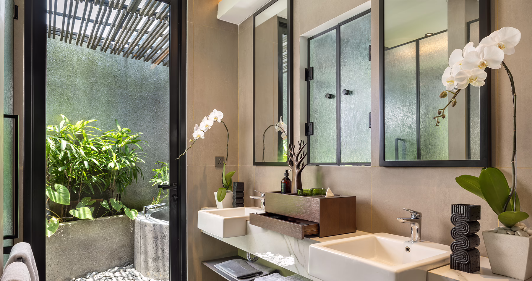 Contemporary bathroom at Quenzo Beach Resort Padangbai featuring twin washbasins, marble countertops, white orchids and a glass door overlooking a private tropical garden.