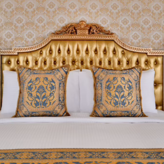 An opulent bedroom at Raffles The Palm Dubai, featuring a gold tufted headboard, ornate furnishings, patterned wallpaper and blue-and-gold cushions.