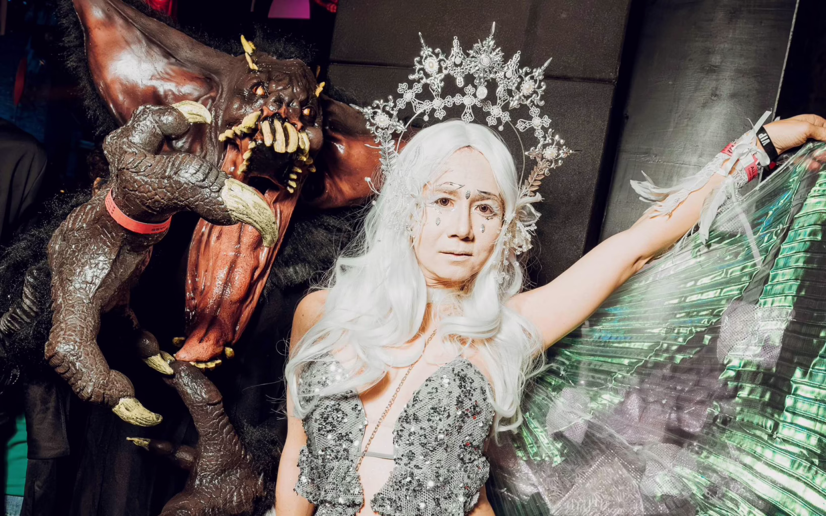 Two elaborately costumed Halloween revellers pose together, one dressed as a horned monster with sharp claws and fangs, the other wearing a silver sequinned outfit, white wig and ornate headpiece.