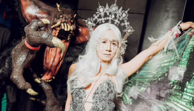 Two elaborately costumed Halloween revellers pose together, one dressed as a horned monster with sharp claws and fangs, the other wearing a silver sequinned outfit, white wig and ornate headpiece.