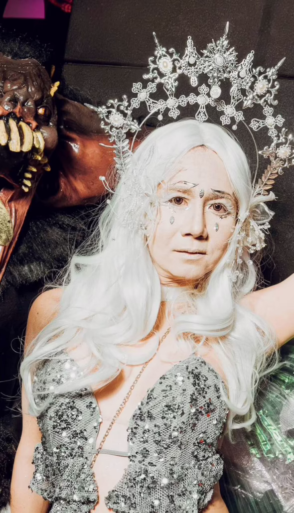 Two elaborately costumed Halloween revellers pose together, one dressed as a horned monster with sharp claws and fangs, the other wearing a silver sequinned outfit, white wig and ornate headpiece.