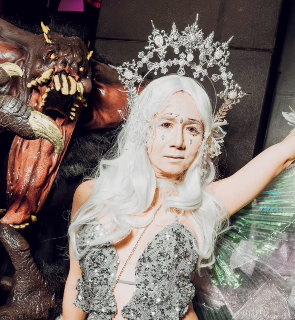 Two elaborately costumed Halloween revellers pose together, one dressed as a horned monster with sharp claws and fangs, the other wearing a silver sequinned outfit, white wig and ornate headpiece.