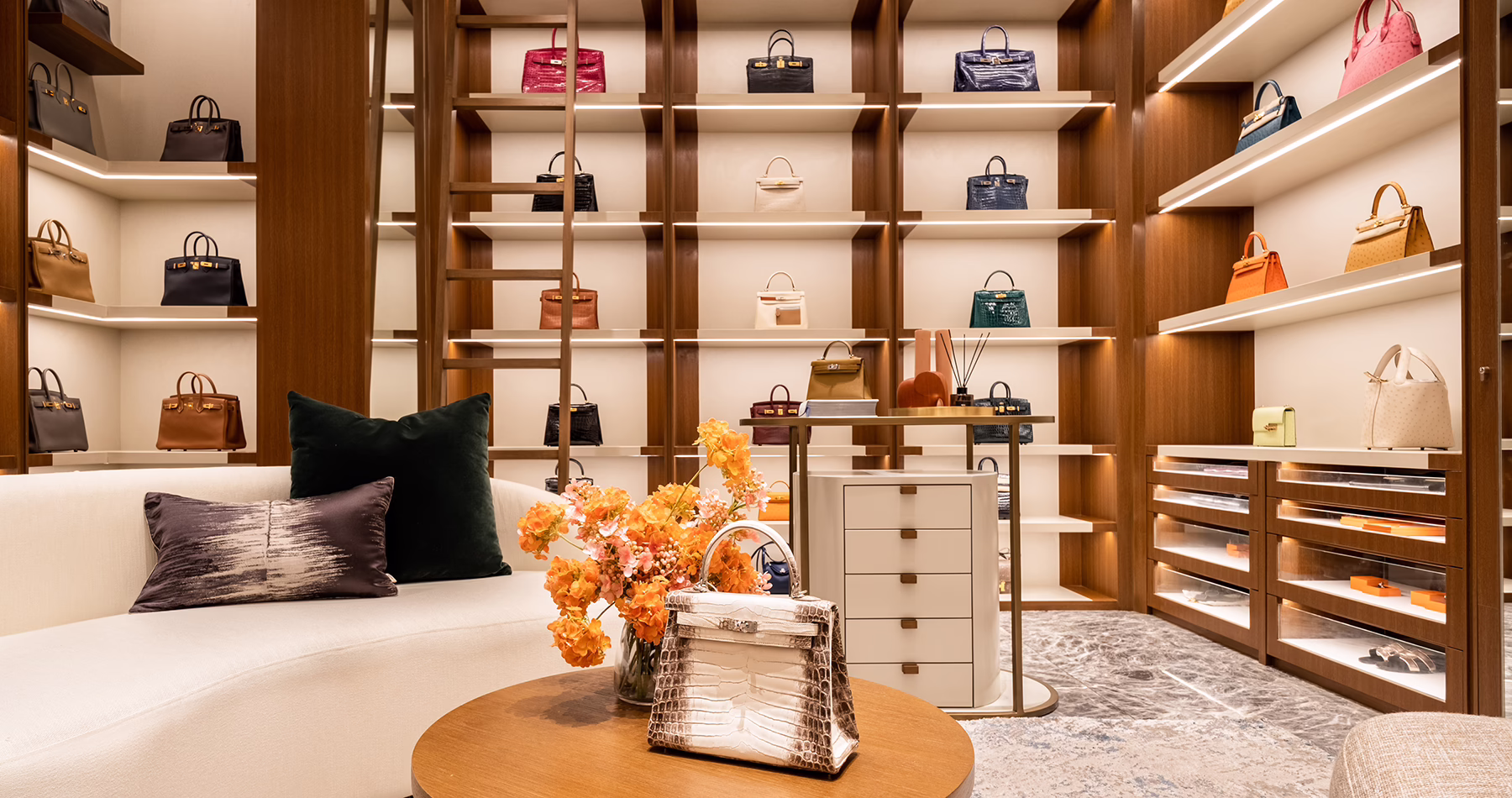 An elegantly furnished showroom at Belle&Kate ROOM in Jakarta, featuring illuminated shelves of Hermès handbags, plush seating and a central display table.