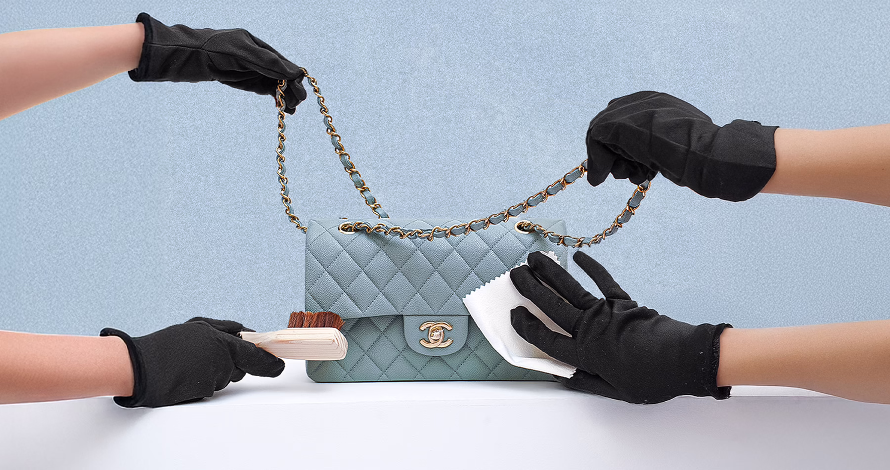 Black-gloved hands carefully clean a blue quilted Chanel handbag using a soft brush and polishing cloth.
