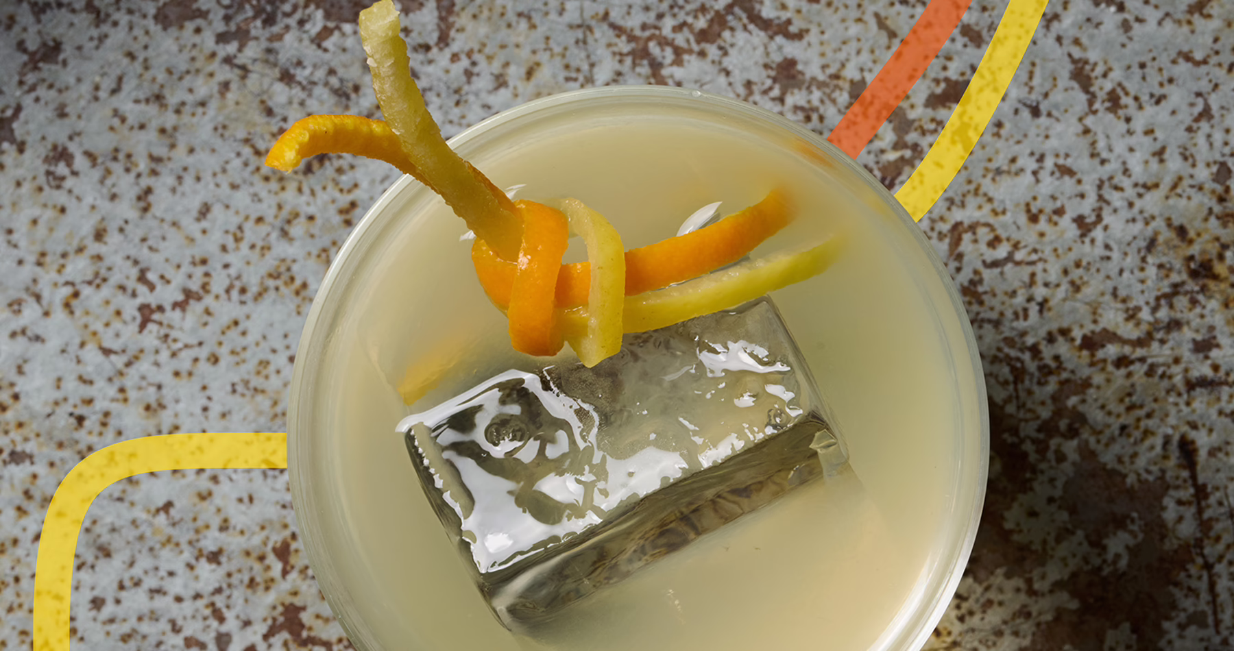 Overhead view of Rita’s Afogilo cocktail, a pale yellow drink served over a large ice cube and garnished with citrus peel.