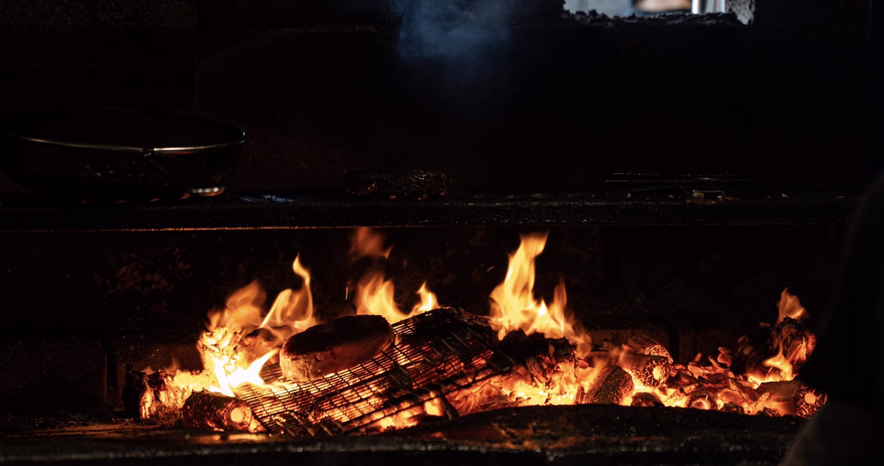Glowing charcoal and burning logs beneath a metal grill inside Firewood's kitchen in Penang.