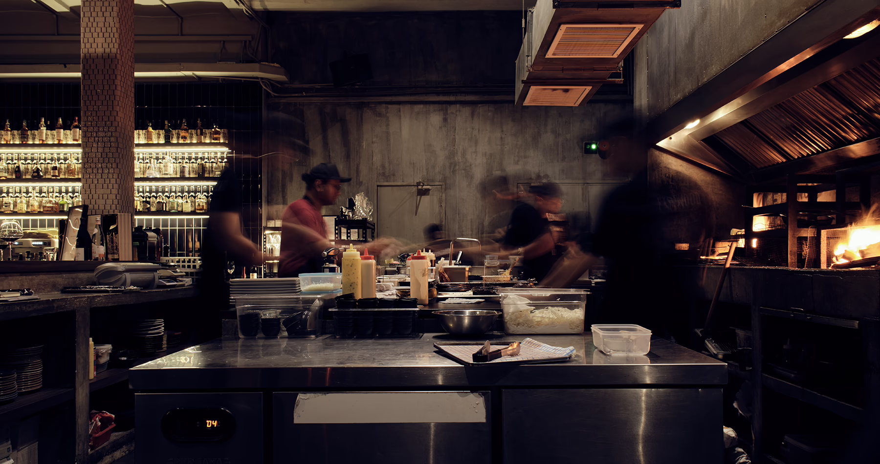Chefs working in Firewood's open kitchen in Penang, with a charcoal-fired grill on one side and an illuminated bar on the other.