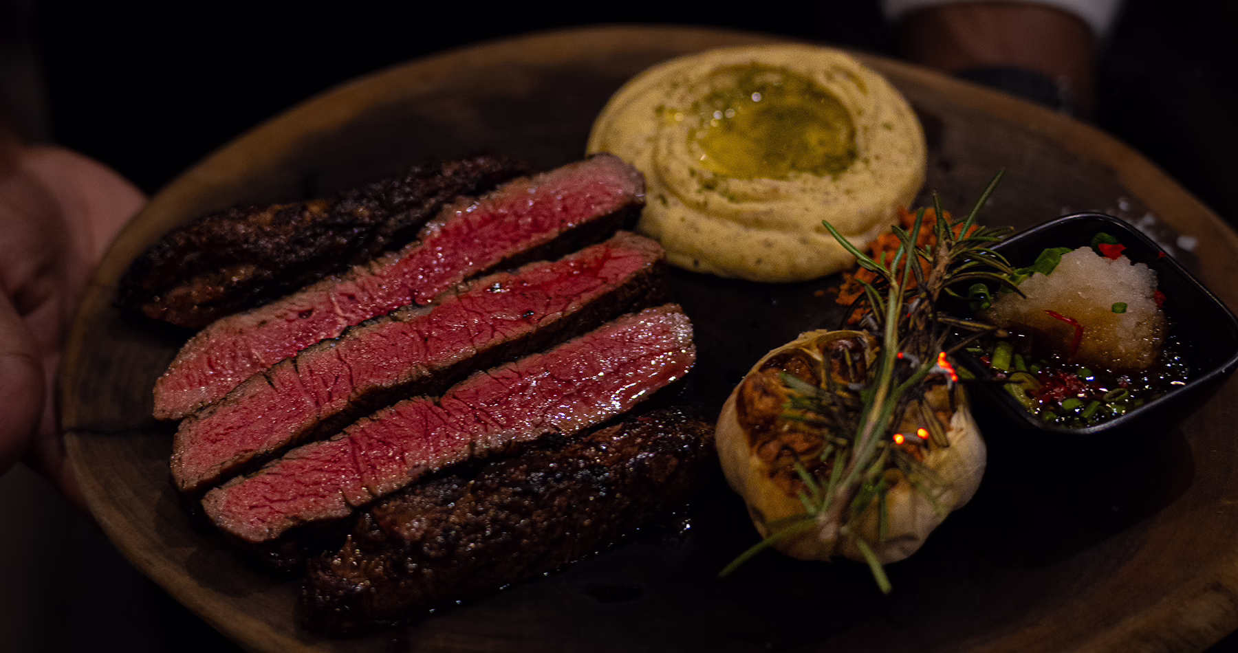 Sliced medium-rare steak served on a wooden platter with roasted garlic, mashed potatoes, rosemary and dipping sauce.