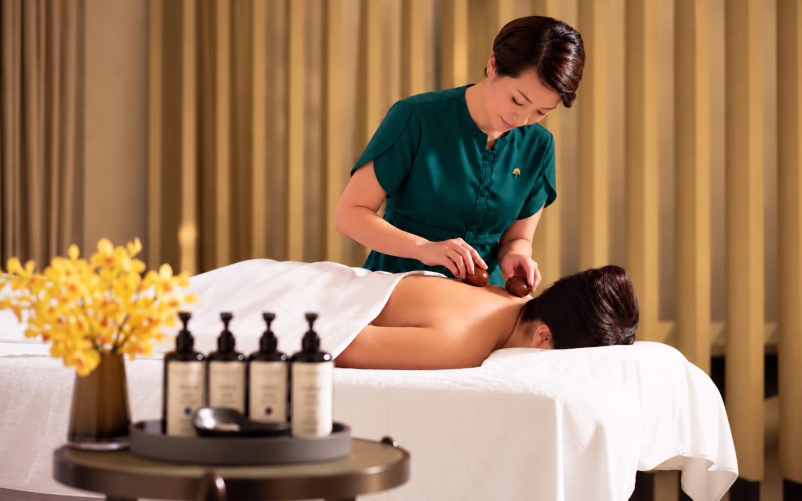 A therapist at Raffles Spa, Singapore, uses smooth gemstone crystals to massage a guest's upper back and shoulders as she lies face down on a treatment bed.