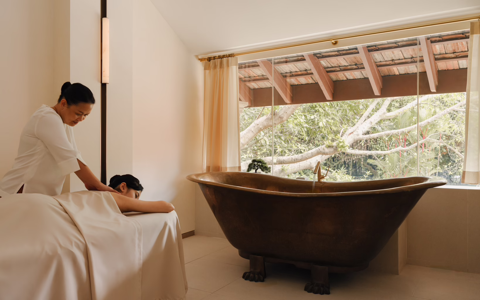 A therapist massages a guest lying face down in a treatment room at Raffles Sentosa Spa, featuring a freestanding bathtub overlooking lush tropical greenery.