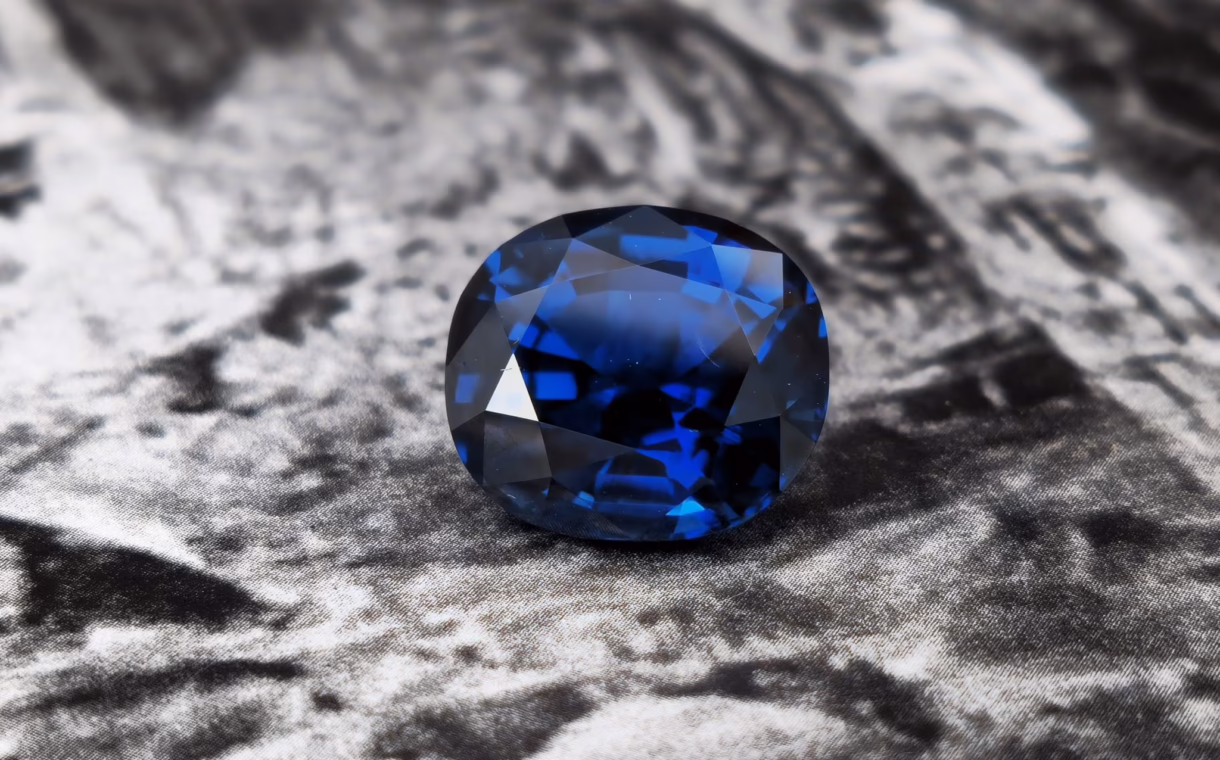 A deep blue, faceted sapphire gemstone displayed against a textured grey background.