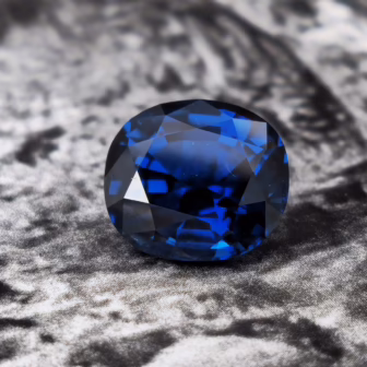 A deep blue, faceted sapphire gemstone displayed against a textured grey background.