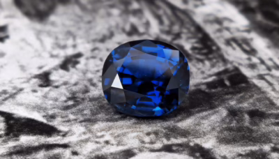 A deep blue, faceted sapphire gemstone displayed against a textured grey background.