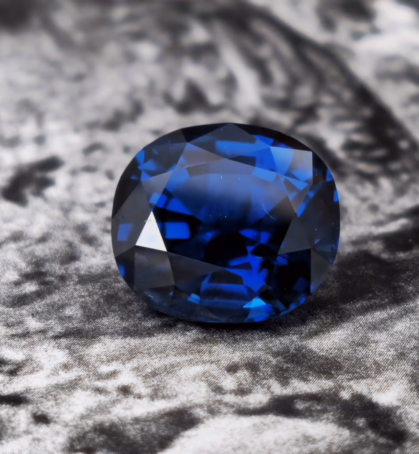 A deep blue, faceted sapphire gemstone displayed against a textured grey background.