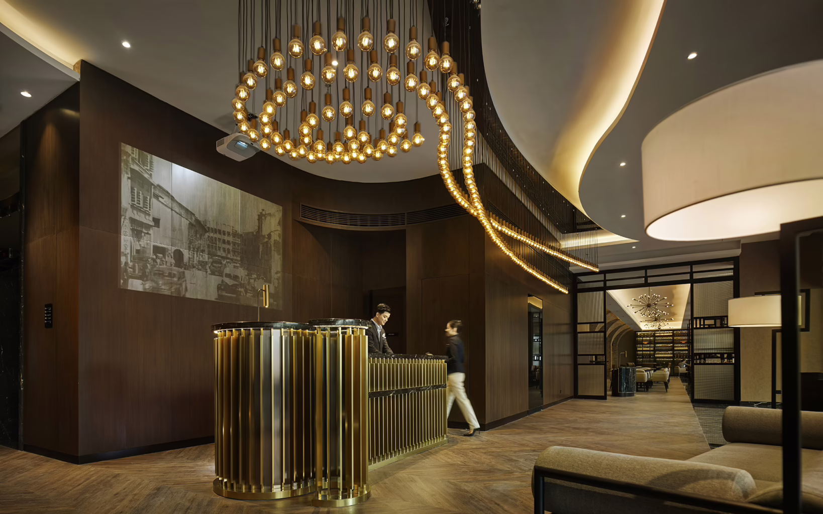 Contemporary lobby of Hotel Stripes Kuala Lumpur, featuring a gold-accented reception desk, sculptural pendant lighting, dark timber walls and a large black-and-white photograph of historic Kuala Lumpur.