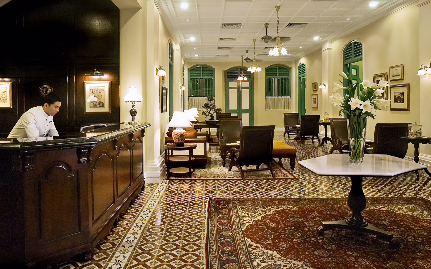 Reception area of The Majestic Malacca, featuring a dark wooden counter, patterned Peranakan floor tiles, green shuttered windows, antique-style furnishings and warm lighting.
